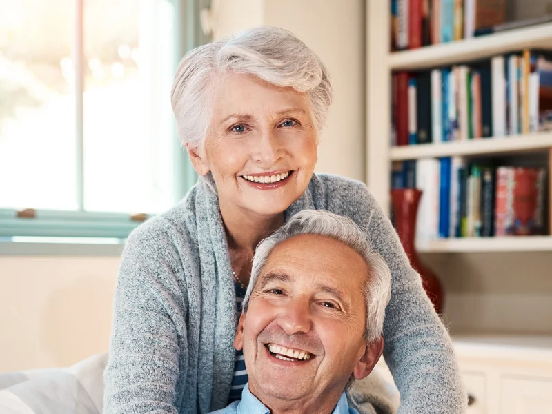 Illustrative photo of a smiling older couple at home, the woman standing behind the sofa with her arms around the man.