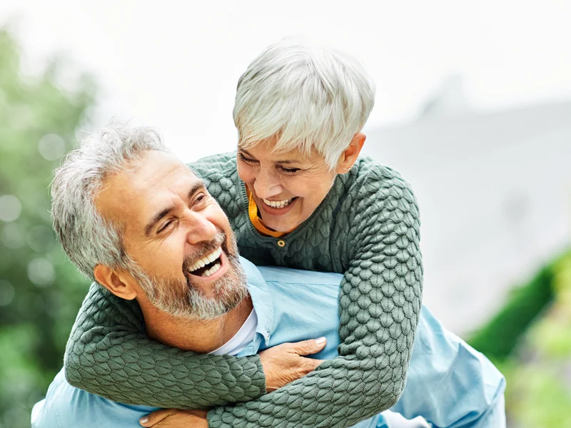 Illustrative photo of a laughing older couple outdoors, the woman riding piggyback on the man's back.