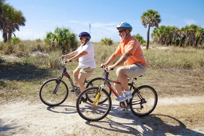 Older couple riding bikes on a sandy trail with palm trees in Florida.